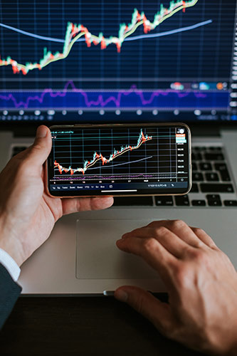 Photo of a hand holding a smartphone with a stock chart, comparing it to a different chart on a large monitor behind the phone