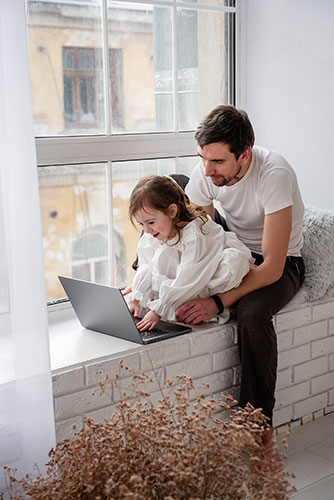 Young father with his little daughter sitting on a window ledge using the laptop to look up disability insurance information