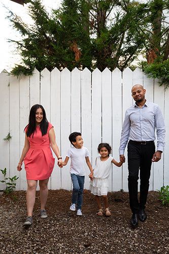 Mom, Dad, and two little kids holding hands in their backyard, standing by a white picket fence