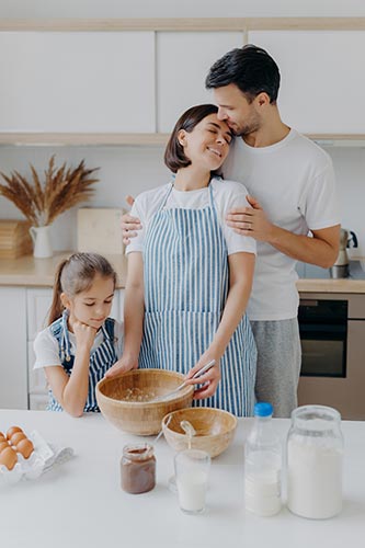 Mom, dad and daughter are cooking together in the kitchen, as the dad kisses the mom on the forehead