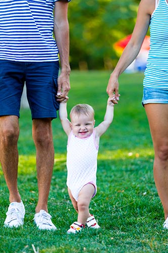 Parents hold their grinning toddler daughter by her hands as she stands in the grass