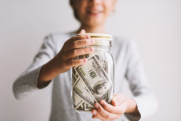 Kid holding a glass jar full of hundred dollar bills, representing the concept of saving