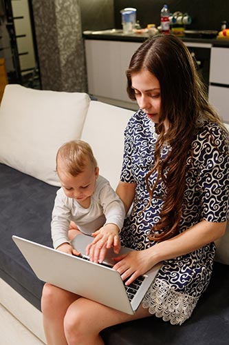 Young mom looking up the benefits of life insurance on her laptop, as her baby plays beside her